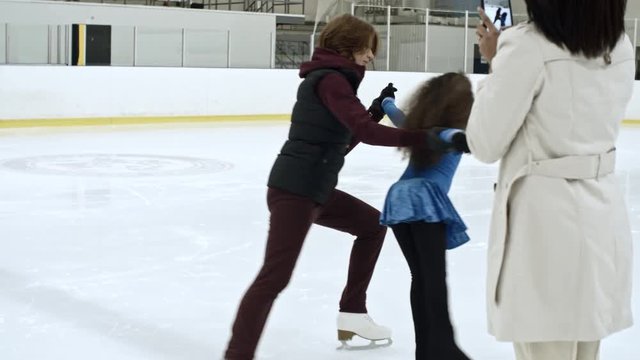 SLOMO of woman recording video with smartphone of her little daughter performing figure skating elements with female coach at indoor ice rink