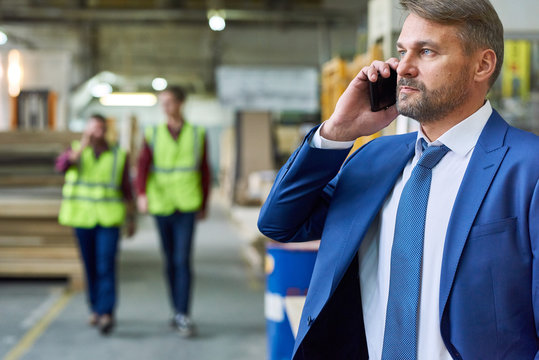 Portrait of handsome mature businessman speaking by phone in factory workshop with two workers in background, copy space