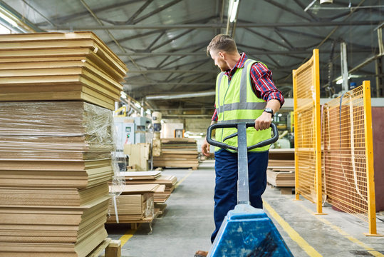 Full length portrait of young man wearing reflective jacket moving wooden boards in factory warehouse, copy space