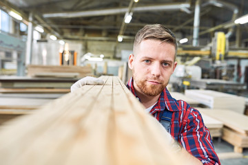 Head and shoulders portrait of handsome young factory worker looking at camera while carrying long wooden board moving material in workshop, copy space