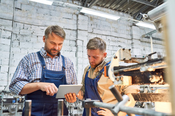 Portrait of two factory workers using modern machine at factory operating it via digital tablet,...