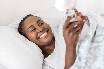 Smiling African woman checking her phone while lying in bed