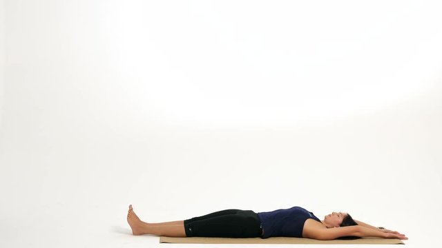 Woman demonstrates stretching exercises and yoga poses on a yoga mat against a white background. She is showing a shoulder stand with legs extended over her head. Side view, profile view.