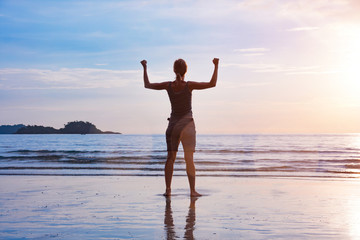 Fototapeta na wymiar Winning and triumph concept. Young woman with stretched up arms standing at idyllic sunset sea beach.