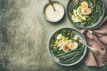 Healthy vegetarian protein rich breakfast bowls. Quinoa, kale, green beans, avocado, egg and creamy tahini dressing bowls over grey concrete background, top view, copy space. Clean eating food concept