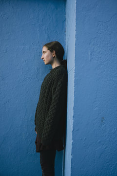 Portrait of teenage girl with brown hair in ponytail wearing black knitted jumper, leaning against blue wall. 