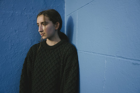Portrait of teenage girl with brown hair in ponytail wearing black knitted jumper, leaning against blue wall.