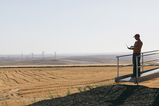 A Wind Farm Technician Standing And Using A Laptop At The Base Of A Turbine On A Wind Farm In Open Countryside At Palouse. 