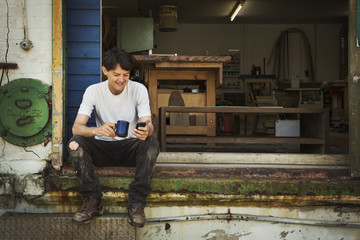 A man in a teeshirt having a tea break, sitting on the steps of a workshop checking his phone. 