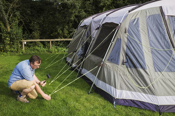 A man using a mallet to hammer in the tent pegs and fix the guy ropes on a tent at a camp site. 