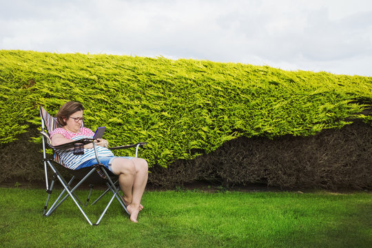 Woman seated in a camping chair in a sheltered spot by a hedge, using a digital tablet.