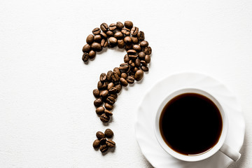 Question mark created from brown beans with cup on the white table. Harvest of coffee in different countries. Choice of the best sort and quality coffee. Empty place for a text.