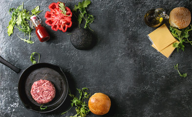 Ingredients for cooking hamburger. Meat beef burger in pan, cheese, ketchup sauce, tomato, black and white buns, arugula salad over dark texture background. Top view with space. Homemade fast food