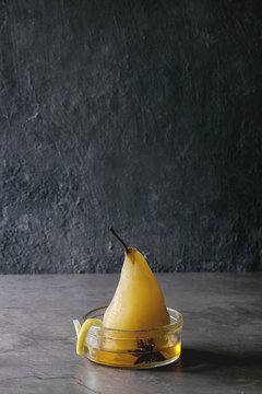 Traditional dessert poached pear in white wine served in glass bowl with syrup and lemon zest over gray texture table.