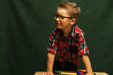 Funny positive boy, an elementary school student wearing glasses and stylish shirt is sitting near wooden school desk with office supplies on dark background