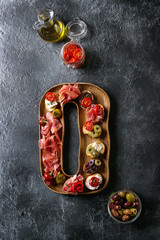 Tapas or bruschetta variety. Crusty bread with ham prosciutto, sun dried tomatoes, olive oil, olives, pepper on decorative wooden plate over dark texture background. Top view with space