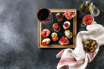 Tapas or bruschetta variety. Bread with ham prosciutto, sun dried tomatoes, olive oil, olives, pepper on slate wood serving board with glass of red wine over dark background. Top view with space