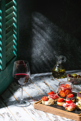 Tapas or bruschetta variety. Bread with ham prosciutto, sun dried tomatoes, olive oil, olives, pepper on slate wood board with glass of red wine over white table. Light via old jalousie. Rustic style.