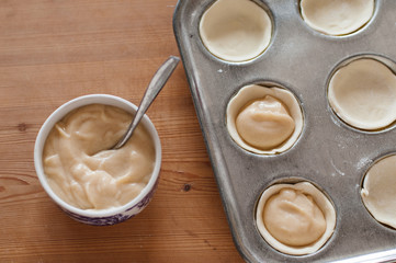 Close up of bowl filled with vanilla, cinnamon egg custard next to baking muffin pan filled with unbaked Pasteis de Nata or Pastel de Belem, a traditional Portuguese egg custard pastry dish