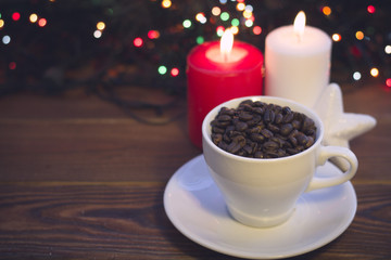 A romantic festive still life with a white cup filled with coffee beans, glitter white star and two burning candles on a rustic wooden table. Dark blurred bokeh background