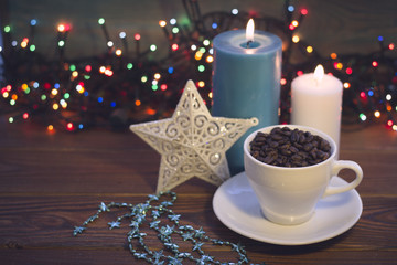 A romantic festive still life with a white cup filled with coffee beans, a chain of blue star shaped beads, a white patterned toy star and blue and white burning candles on a rustic wooden table. Dark