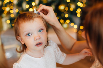 Cheerful happy woman putting little crown in daughter's hair near christmas tree. New Year's interior.