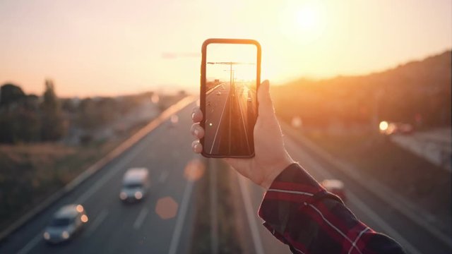 Cinemagraph of social media influencer of mobile photographer hold smartphone with high resolution screen, make video of busy evening traffic on highway in sunset. concept commuting and social media