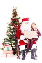 Santa Claus reading a book for cute little girl sitting on his lap near Christmas tree