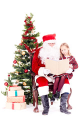 Fototapeta na wymiar Santa Claus reading a book for cute little girl sitting on his lap near Christmas tree