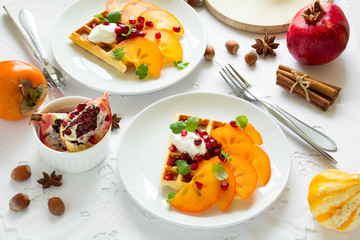 Plates of Belgian waffles with persimmon, pomegranate seeds and sour cream on white table cloth