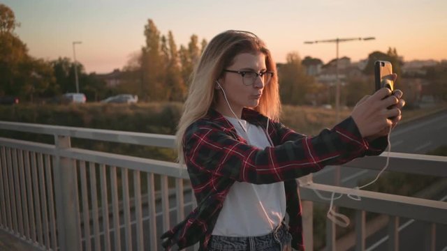 Beautiful blonde woman, hipster cool student or social media influencer or blogger, walks on busy street at sunset and makes selfie photographs on mobile smartphone, blows a kiss to camera