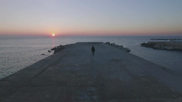 Aerial footage of drone fly over pier or concrete boardwalk at empty beach at sunset or sunrise, lonely figure of hipster man in sailor outfit, winter coat and sweater, concept adventures and explore