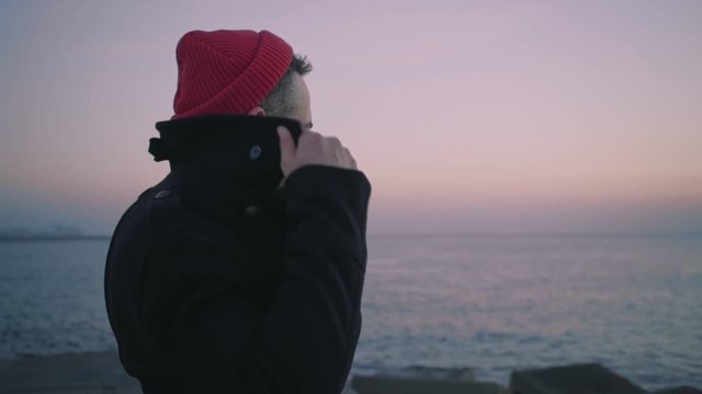 Slow motion shot of romantic and authentic icelandic man, in adventurer outfit, black coat and red sailor or fisherman hat. look at sunset or sunrise, lost in thoughts, or dreams of future