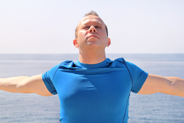 Athletic fit runner doing stretching exercise, preparing for morning workout on a seashore. A handsome fitness athlete doing a stretching routine in the sun. Blue sky and the sea in background.