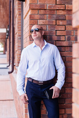 Fototapeta premium bald businessman staing at red brick wall building. shirt dark blue trousers, sunglasses. outdoor spring, summer