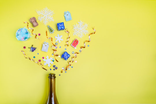 Part of champagne party bottle with confetti of gifts, sequins, ribbons, snowflakes on bright yellow background. Flat lay. Celebrate new year concept. Selective focus, space for text
