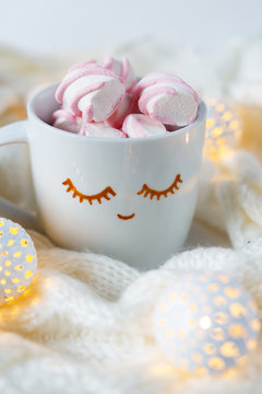 White cup with cocoa and marshmallow with painted eyes in a cozy scarf on a light background with copy space
