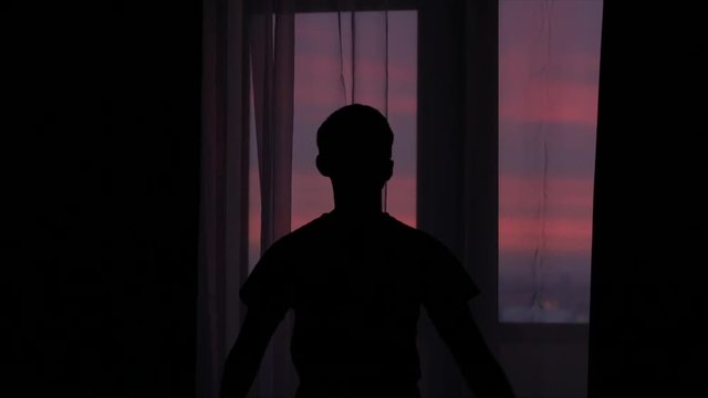 silhouette of a young man opening curtains against a window with a beautiful sunset