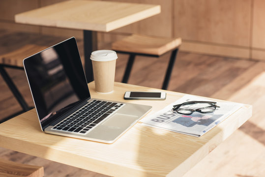 Laptop With Blank Screen, Smartphone And Business Newspaper On Table In Coffee Shop