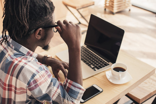 african american man using laptop and smartphone with blank screens in coffee shop