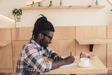 Fototapeta na wymiar african american man writing in notepad during coffee break in cafe