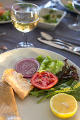 Delicious seafood meal baked fish with vegetables and lemon on a plate in the restaurant. Glass of wine, forks and knives on the table, all prepared for celebration evening. Dim sunset light, outdoors