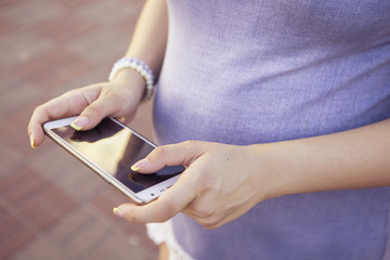 Beautiful brunette caucasian pregnant woman on summer walk outdoors, holding her belly in a soft, sensual, maternity way. In casual jeans dress and bride hairstyle typing message on phone. Copy space