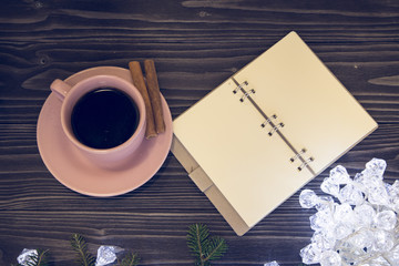 A dark wooden Christmas background with white fairy lights . A cup of coffee with cinnamon sticks on a saucer and a blank notebook on a table. Top view. Copyspace