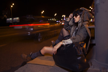 Fototapeta na wymiar Beautiful brunette fashion casual stylish woman (looks like homeless) outdoor at night in city lights, with a bag of clothes, near the road