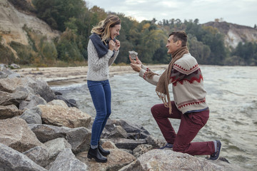 Happy thoughtful couple standing on a rock beach near sea hugging each other in cold foggy cloudy autumn weather. Man make proposal to woman with a huge rock. Copy space