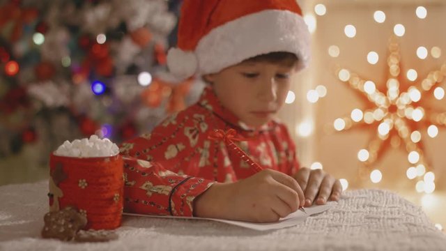 Dreamy boy in pajamas and santa hat writing the letter to Santa with the cup of cocoa with marshmallow
