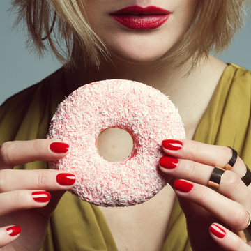 Fashion & Gluttony, Symbol of Woman Concept. Gorgeous model in mustard cocktail dress eating donut over gray background. Perfect skin, make-up, manicure. Golden accessories. Close up. Studio shot