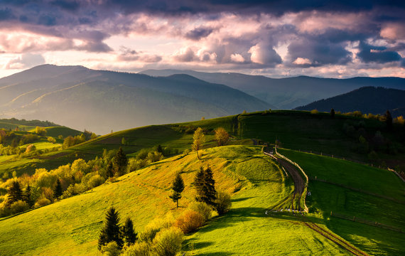country road through green rolling hills at sunset. gorgeous springtime rural landscape in mountains under the sky with pink clouds