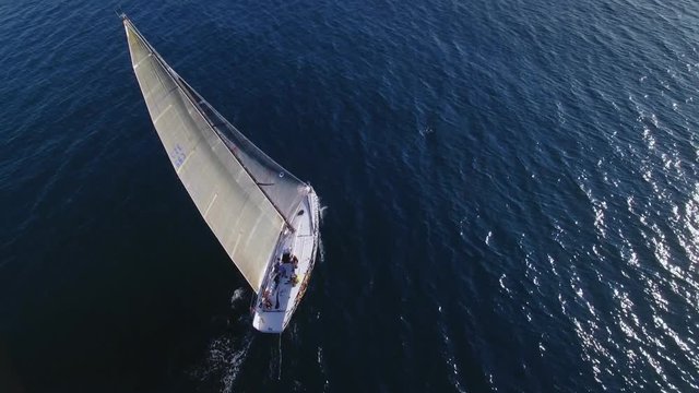 from drone of professional racing competition sailboat with spinnaker and mainsail up, moves on fast nautical marine speed in vast blue deep waters of ocean, white sails in air